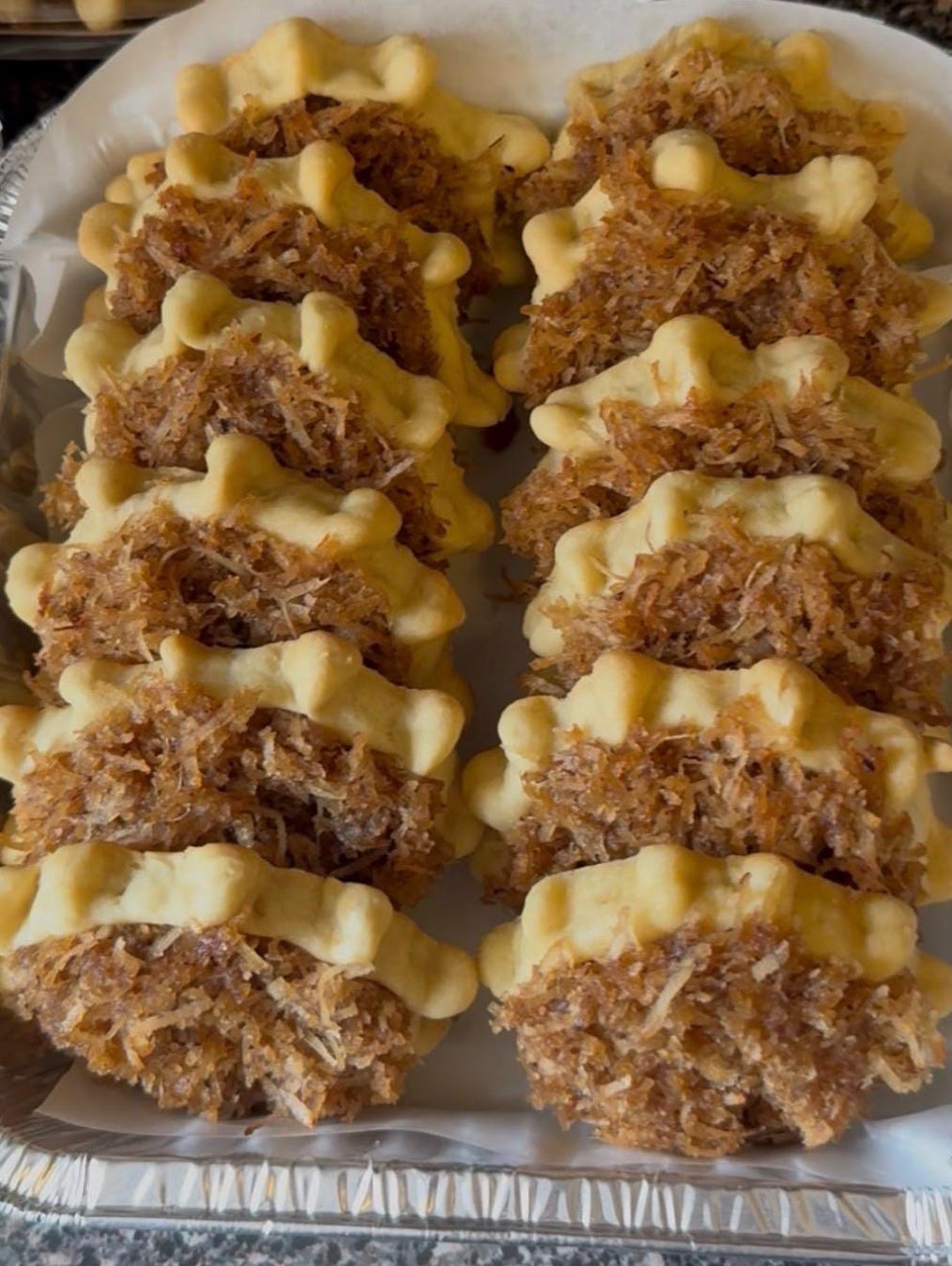 A tray of coconut shrimp paste pockets filled with shredded coconut and topped with a drizzle of yellow sauce, arranged in a foil pan on parchment paper.